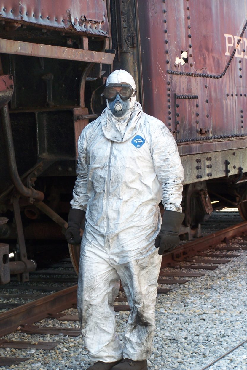 Worker dressed in sandblasting gear posing for a photo in front of an old locomotive