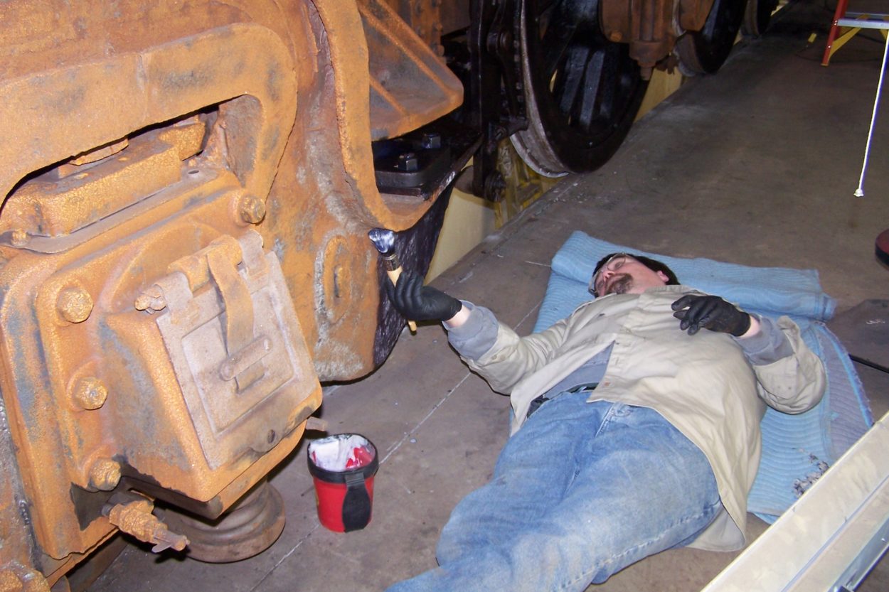 Worker laying on his back while holding a paint brush, applying grease to a locomotive