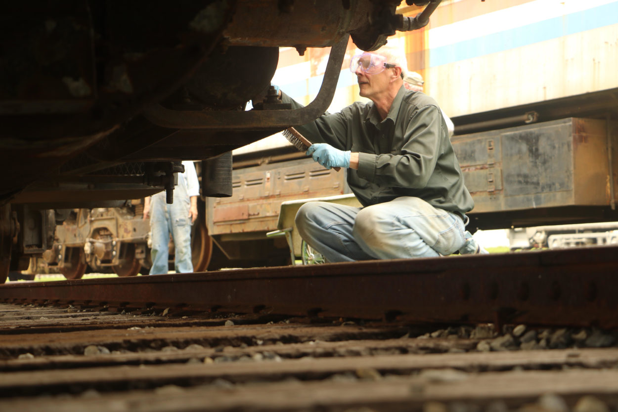Worker using a steel brush to remove rust from the exterior surface of a locomotive